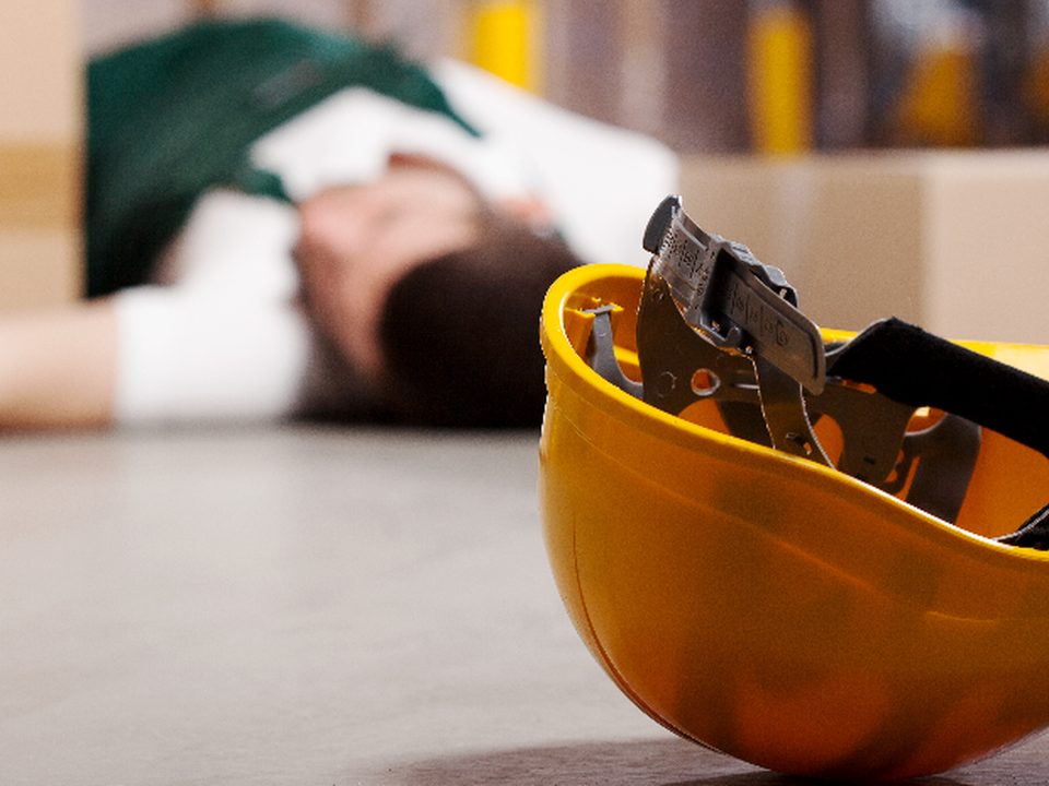 Arbeitsunfall: Schutzhelm am Boden, verletzte Person im Hintergrund